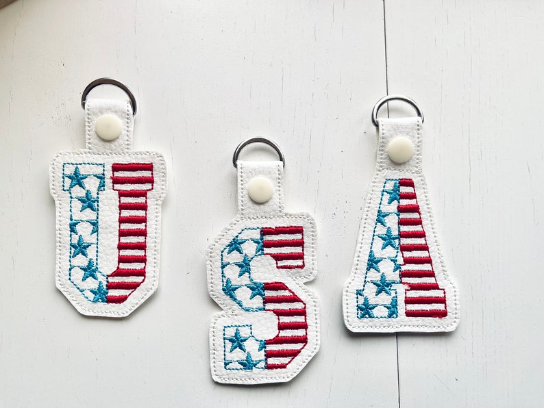 May include: Three keychains with the letters U, S, and A embroidered in red, white, and blue. The letters are filled with a pattern of red and white stripes and blue stars.