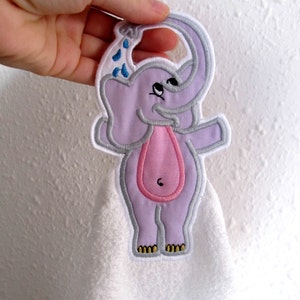 Puede incluir: Un elefante morado con una trompa rosa y gotas de agua azules bordadas en una toalla blanca. El elefante tiene un lazo en la cabeza para colgar.