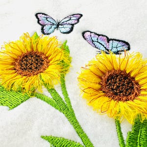 May include: Embroidered design featuring two yellow sunflowers with brown centres and two purple butterflies on a white background.