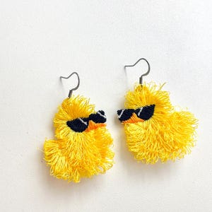 May include: A pair of handmade yellow duck earrings with black sunglasses. The earrings are made of yellow thread and have silver-colored ear wires. The duck earrings are a fun and quirky accessory.