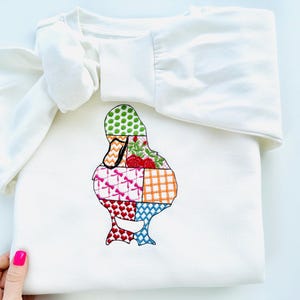 May include: A white long-sleeved shirt with a patchwork duck design embroidered on the front. The duck is made up of different coloured squares with patterns like polka dots, plaid, and hearts.