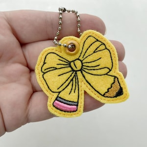 May include: A yellow felt keychain shaped like a bow combined with a pencil. The design features black embroidered outlines, a pink eraser tip, and a metallic ball chain attached through a small yellow grommet at the top of the bow.
