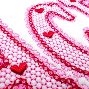 Könnte beinhalten: Nahaufnahme eines weißen Stoffes mit einem rosa und roten Stickmuster. Das Design zeigt ein sich wiederholendes Muster aus kleinen roten Herzen und einen gewellten Rand. Die Stickerei ist detailliert und aufwendig.
