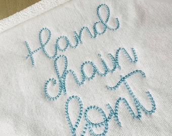 May include: Close-up of a white fabric with the words "Hand chain font" embroidered in light blue thread. The text is in a cursive style, with each letter meticulously stitched. The fabric has a textured appearance.