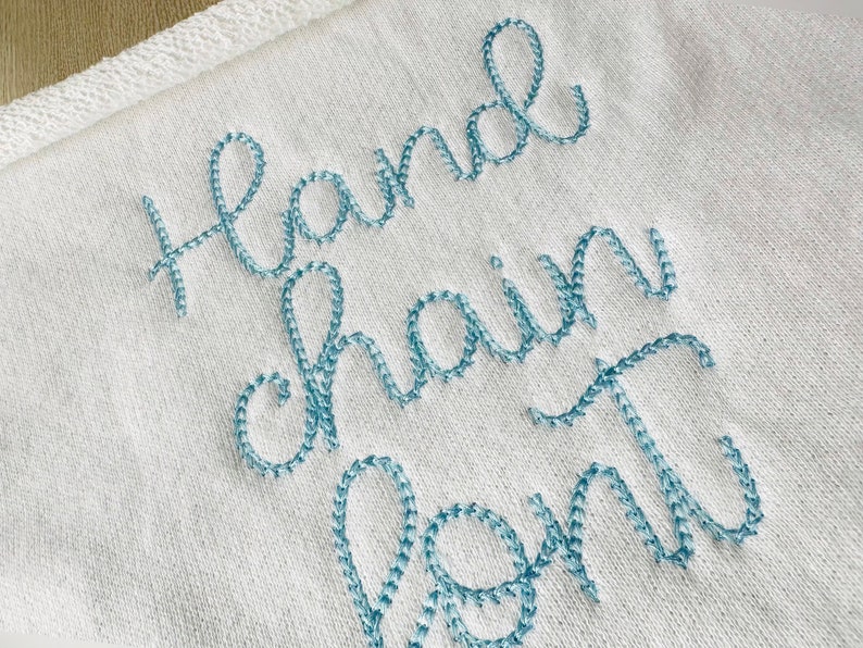 May include: A white towel with the words "hand chain font" embroidered in light blue thread.
