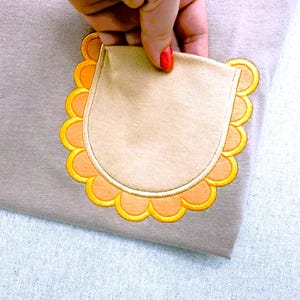 Puede incluir: Una prenda de tela marrón claro que presenta un bolsillo de parche con un borde festoneado. El bolsillo es de un color beige más claro y está bordeado por un ribete bordado amarillo brillante que crea un patrón decorativo de festones redondeados.