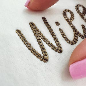 Może przedstawiać: Zbliżenie haftowanego tekstu na białej tkaninie. Tekst, w stylu kursywy, jest wyszyty brązową nicią. Haft ma wygląd łańcuszka. W kadrze widoczny jest różowy paznokieć.