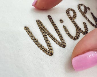 Puede incluir: Primer plano de texto bordado sobre tela blanca. El texto, en estilo cursivo, está cosido con hilo marrón. La costura tiene apariencia de cadena. En el encuadre se ve una uña rosa.
