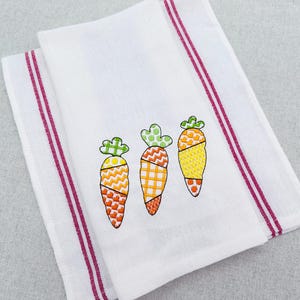 以下が含まれることがあります： オレンジ、黄色、緑色の刺繍された3つのニンジンが付いた白いキッチンタオル。ニンジンにはさまざまな幾何学模様があります。タオルには2本の垂直のバーガンディ色のストライプがあります。