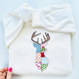 May include: A white sweatshirt with a patchwork deer design embroidered on the front. The deer is made up of different colored squares, including blue, pink, green, and orange.