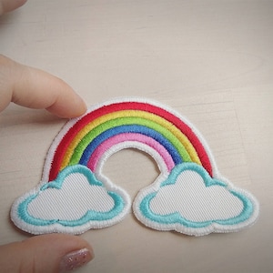 May include: A rainbow-colored felt patch with a red, orange, yellow, green, blue, and purple arc. The rainbow has two white clouds with a light blue outline at each end.
