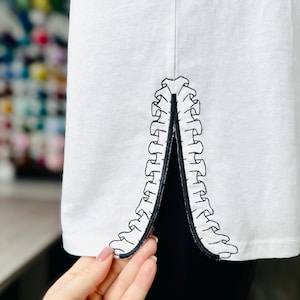 Puede incluir: Camiseta blanca con un diseño bordado en negro de una columna vertebral en la costura lateral.