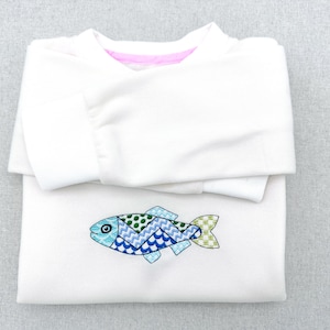 May include: A folded white sweatshirt with a colorful embroidered fish design. The fish features blue, green, and teal patterns. The neckline has a pink trim.