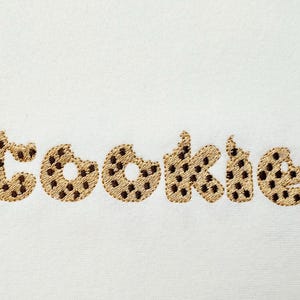 Puede incluir: Palabra bordada "COOKIE" en una fuente lúdica, que se asemeja a galletas con chispas de chocolate. Las letras son de color canela con detalles de chispas de chocolate marrón, sobre un fondo blanco. Un diseño divertido con temática de comida.