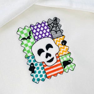 May include: A white skull with black eyes and a smile is centered on a colorful patchwork design. The patchwork includes green, purple, orange, and teal squares with Halloween-themed images like bats and spiders. The design has a zig-zag border.