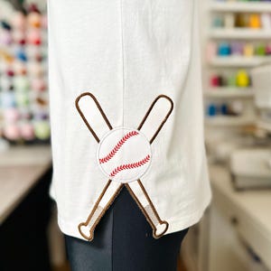 May include: White baseball-themed shirt with an embroidered design. The design features a baseball with red stitching and two crossed bats in brown. The shirt is made of a soft, light-colored fabric.