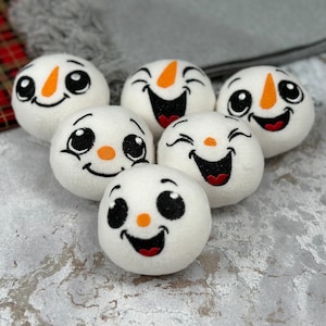 May include: Six white snowman face decorations with embroidered black eyes, smiles, and orange noses. The snowmen have a plush texture and are arranged on a textured grey surface. Festive holiday decor.