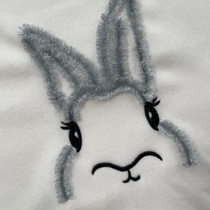 May include: A white fabric with a gray embroidered bunny face. The bunny has black eyes and a black nose.