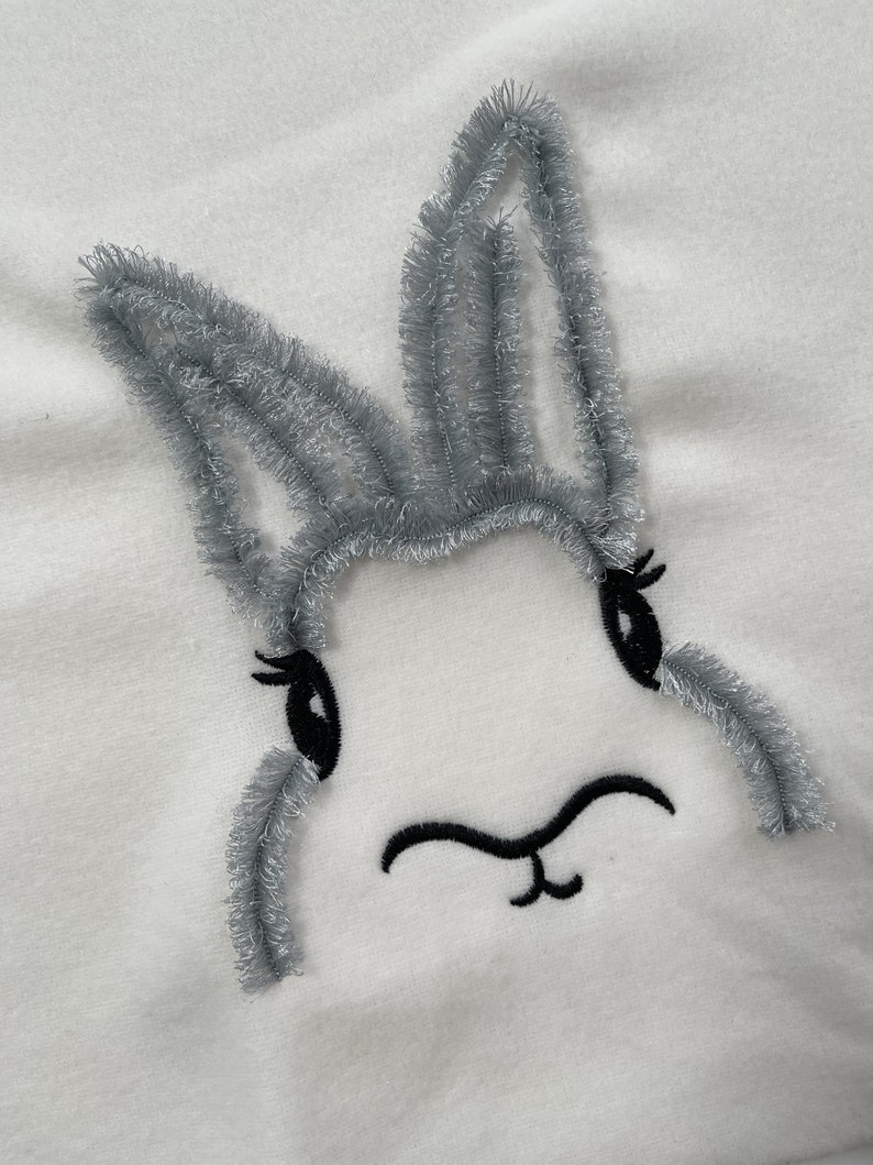 May include: A white fabric with a gray embroidered bunny face. The bunny has black eyes and a black nose.