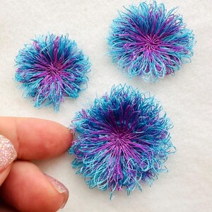 May include: Three handmade blue and purple fabric flowers. The flowers are made of thin, layered fabric and have a fluffy, pom-pom like appearance.
