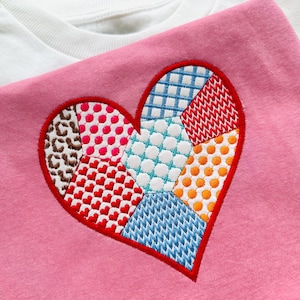 May include: A pink t-shirt with a red heart-shaped patch made of different colored fabric squares. The squares are stitched together in a patchwork pattern and include colors like red, blue, orange, white, and brown.