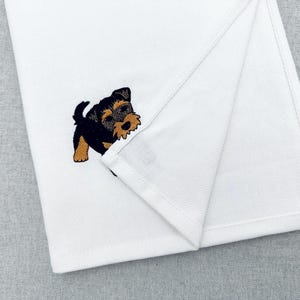 以下が含まれることがあります： ヨークシャーテリアの子犬が刺繍された白い折り畳みタオル。子犬は黒、タン、茶色です。タオルは柔らかい白い生地で作られています。