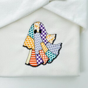 May include: A white fabric item with a colorful patchwork ghost applique. The ghost design features various patterns and colors, including orange stripes, purple squares, and yellow hearts. The ghost has black eyes and a black lace trim.
