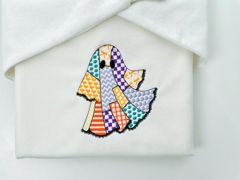 May include: A white fabric item with a colorful patchwork ghost applique. The ghost design features various patterns and colors, including orange stripes, purple squares, and yellow hearts. The ghost has black eyes and a black lace trim.