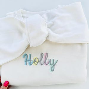 Könnte beinhalten: Cremefarbenes Sweatshirt mit dem Namen "Holly" in Pastellfarben gestickt. Das Sweatshirt hat eine große, dekorative Schleife am Ausschnitt. Das Bild ist aus der Vogelperspektive aufgenommen.