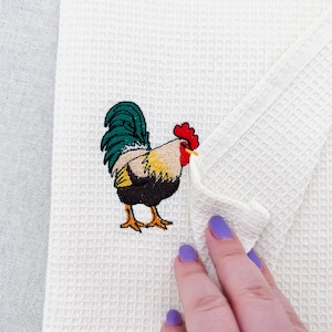 Op de afbeelding: Witte keukenhanddoek met wafelweefsel en een geborduurd hanenontwerp. De haan heeft een groene staart, een rode kam, een gele snavel en een zwart en geel lichaam. Een hand met paarse nagellak houdt de handdoek vast.