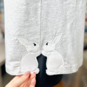 Può includere: T-shirt grigio chiaro con due applicazioni di conigli bianchi vicino all'orlo. I conigli si guardano, con occhi neri e un design semplice. La maglietta è realizzata in un materiale morbido, probabilmente cotone.