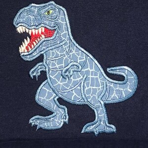 K&ouml;nnte beinhalten: Eine blaue gestickte Dinosaurier-Applikation auf einem dunkelblauen Stoffhintergrund. Der Dinosaurier blickt nach rechts und hat eine rissige Textur.