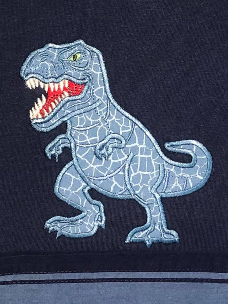 K&ouml;nnte beinhalten: Eine blaue gestickte Dinosaurier-Applikation auf einem dunkelblauen Stoffhintergrund. Der Dinosaurier blickt nach rechts und hat eine rissige Textur.