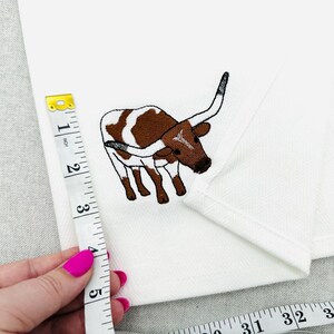 May include: White fabric with an embroidered brown and white longhorn steer. A measuring tape shows the fabric is approximately 5 inches long. The steer has large horns and is facing left.