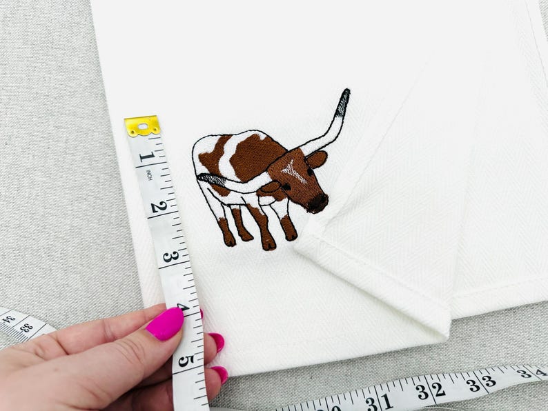 May include: White fabric with an embroidered brown and white longhorn steer. A measuring tape shows the fabric is approximately 5 inches long. The steer has large horns and is facing left.
