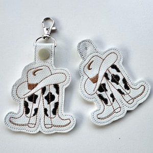 Könnte beinhalten: Weißer Leder-Schlüsselanhänger mit Kuhmuster. Der Schlüsselanhänger zeigt ein Paar Cowboystiefel und einen Cowboyhut.