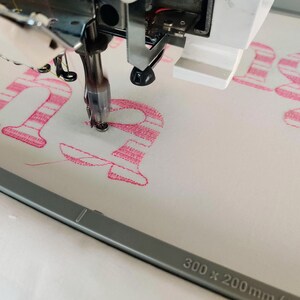 Pode incluir: Close-up de um desenho de bordado listrado rosa e branco sendo costurado por uma m&aacute;quina de costura. O desenho diz "ha" e est&aacute; sendo costurado em um tecido branco. A m&aacute;quina de costura &eacute; um modelo Brother e o aro &eacute; de 300 x 200 mm.