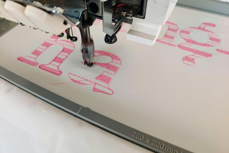 Pode incluir: Close-up de um desenho de bordado listrado rosa e branco sendo costurado por uma m&aacute;quina de costura. O desenho diz "ha" e est&aacute; sendo costurado em um tecido branco. A m&aacute;quina de costura &eacute; um modelo Brother e o aro &eacute; de 300 x 200 mm.