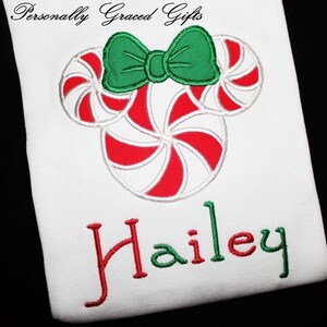 May include: A white t-shirt with a red and white candy cane design in the shape of Mickey Mouse ears with a green bow. The name "Hailey" is embroidered in red and green below the candy cane design.