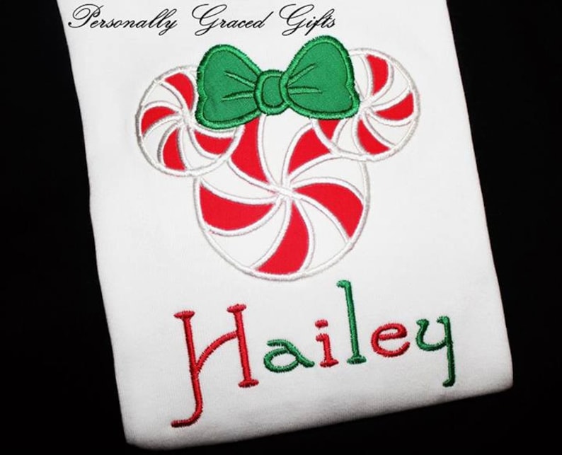 May include: A white t-shirt with a red and white candy cane design in the shape of Mickey Mouse ears with a green bow. The name "Hailey" is embroidered in red and green below the candy cane design.