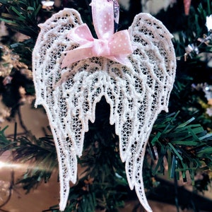 Peut inclure: Ornement en forme d'ailes d'ange en dentelle blanche délicate. Les ailes sont ornées d'un nœud et d'un ruban rose clair à pois. L'ornement est suspendu à un sapin de Noël, ajoutant une touche d'élégance aux décorations de fêtes.