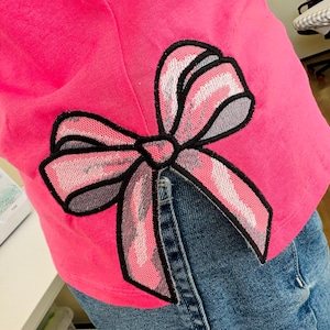 Op de afbeelding: Een roze shirt met een grote geborduurde strik op de rug. De strik is gemaakt van roze, wit en grijs stof en heeft een zwarte omtrek.