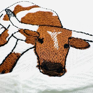 May include: Close-up of an embroidered image of a longhorn steer. The steer has brown and white patches, with a white "T" on its forehead. The embroidery is detailed with black outlines, set against a white background.