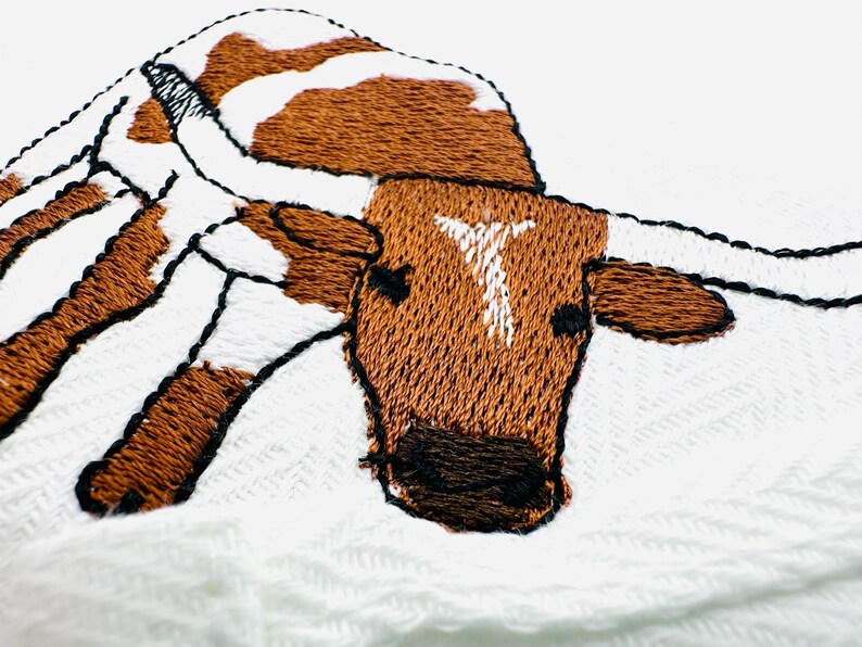 May include: Close-up of an embroidered image of a longhorn steer. The steer has brown and white patches, with a white "T" on its forehead. The embroidery is detailed with black outlines, set against a white background.