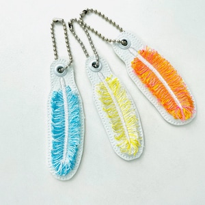 Puede incluir: Tres llaveros blancos con diseños de plumas bordadas en azul, amarillo y naranja. Cada llavero tiene una cadena de bolas plateada y un cierre metálico. Las plumas están detalladas con hilo texturizado, creando un accesorio vibrante.