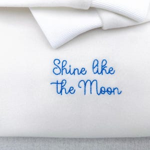 May include: Close-up of a white fabric with the embroidered phrase "Shine like the Moon" in blue cursive script. A white bow is visible at the top. The fabric has a soft texture.
