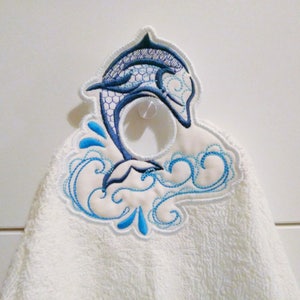 May include: White hand towel with a blue and white embroidered dolphin jumping out of the water. The dolphin has a hole in the middle for hanging.
