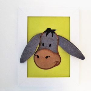 May include: A framed felt artwork of a grey donkey with brown eyes and a brown nose. The donkey is on a yellow background.