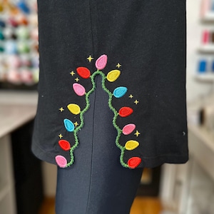 May include: Black garment featuring an embroidered design of festive Christmas lights. The lights are in red, pink, yellow, blue, and green, with small gold stars. The design is on the lower edge of the garment.