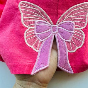 Könnte beinhalten: Pinkfarbener Stoff mit einer weißen und lilafarbenen Schmetterlingsapplikation mit einer Schleife in der Mitte.